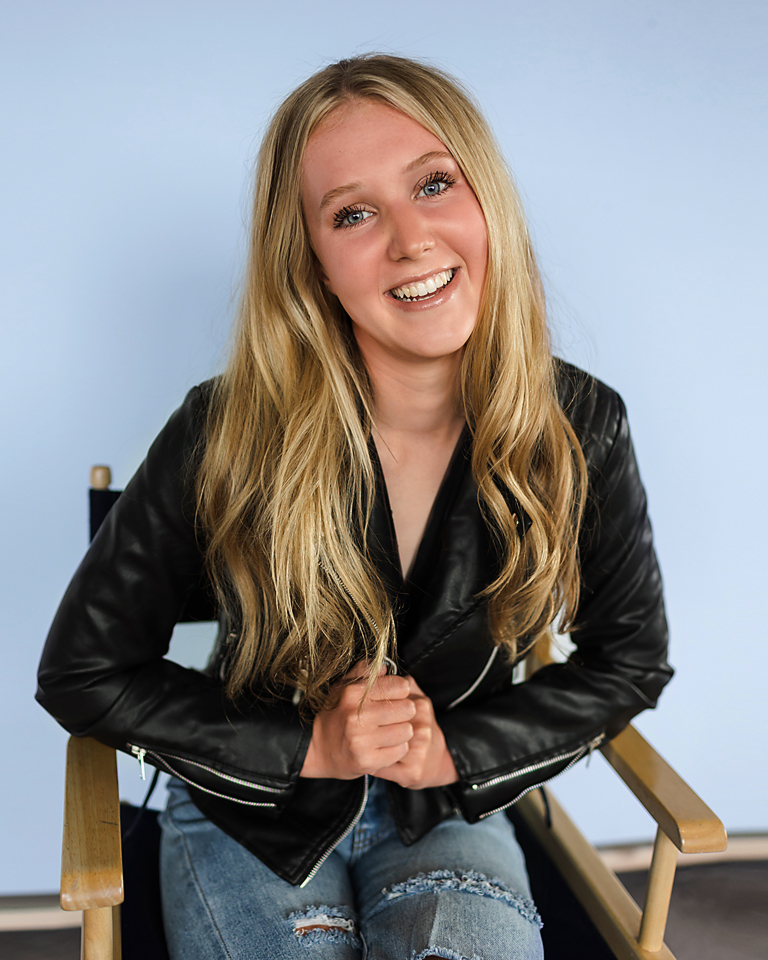 senior sitting in directors chair with a baby blue background in photography studio 